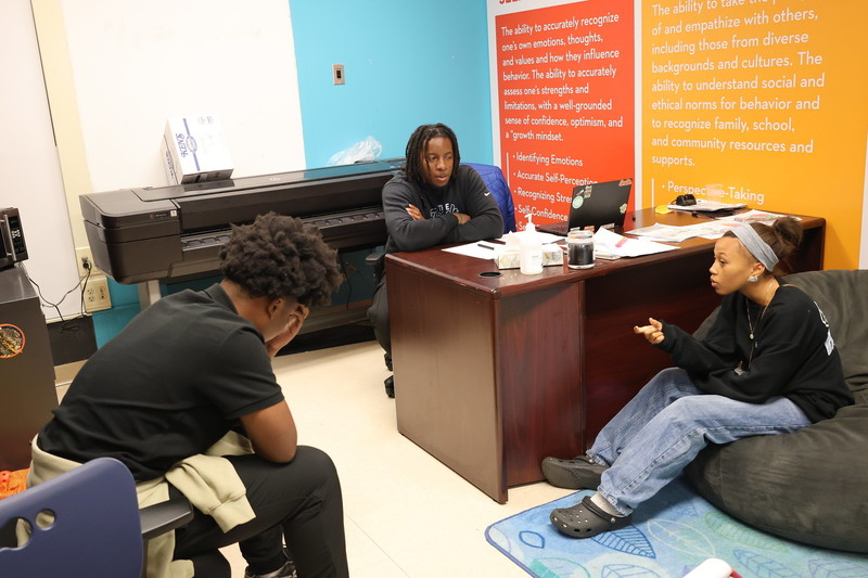 Two students and one teacher sitting in the teacher's office having a casual conversation