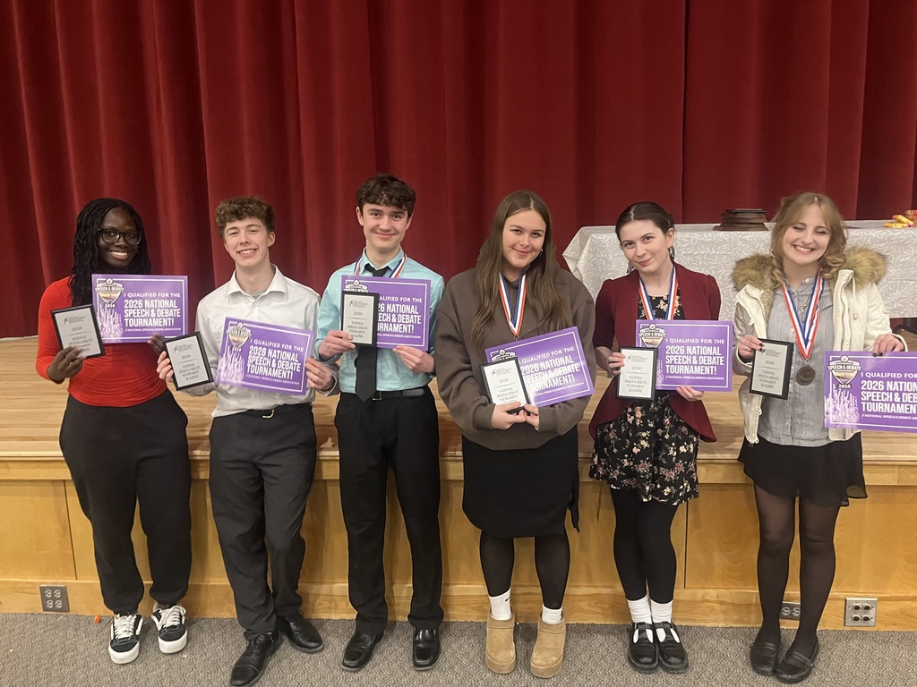 Six individuals stand on a stage with medals and awards, holding purple certificates. A red curtain is behind them.