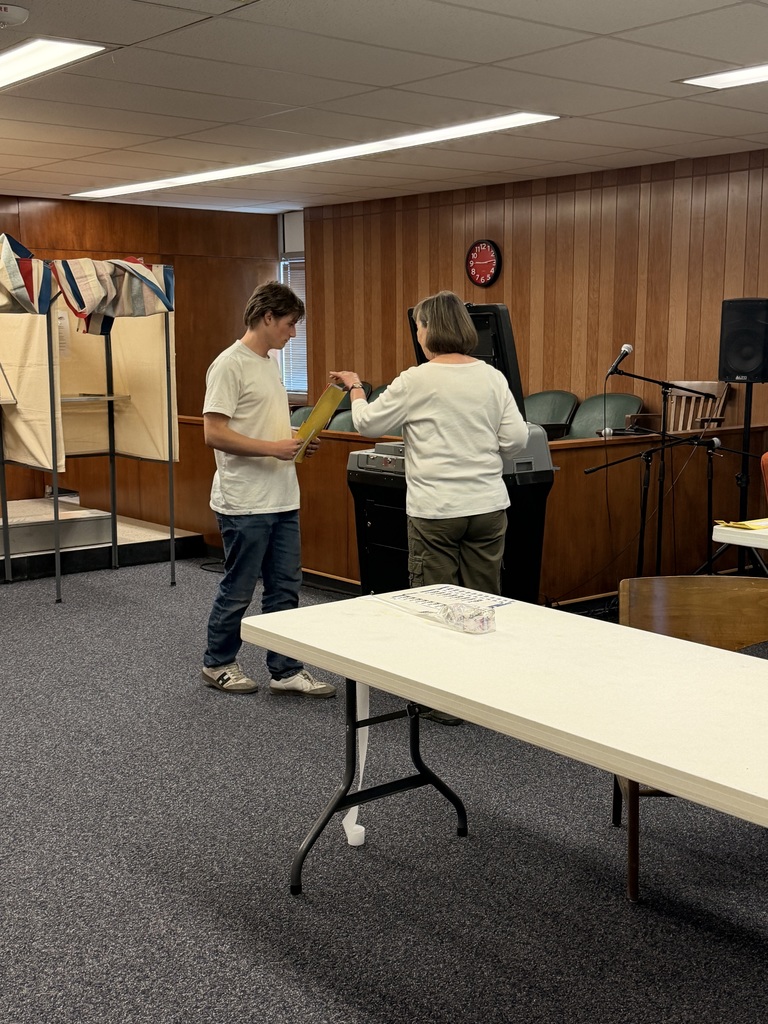 A student in a white t-shirt and a staff member in a white long-sleeve shirt stand together in a wood-paneled room, looking at a yellow document near a podium.