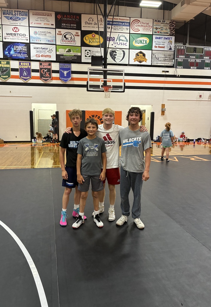 Alt Text: Four young male athletes standing side-by-side and smiling on a black wrestling mat inside a gymnasium. They are dressed in athletic t-shirts and shorts, with one shirt featuring the word "Crawford" and another displaying "Wildcats." The gym floor and other students are visible in the blurred background.