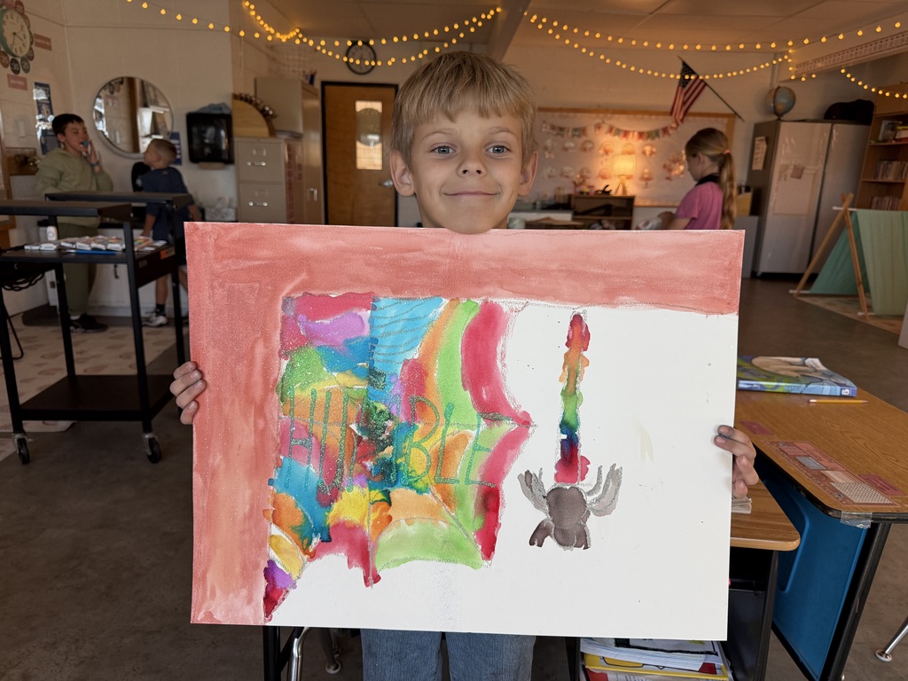 A young student smiling while proudly holding up a colorful watercolor painting of a spiderweb, featuring the word "HUMBLE" in green letters and a spider dangling from a thread.