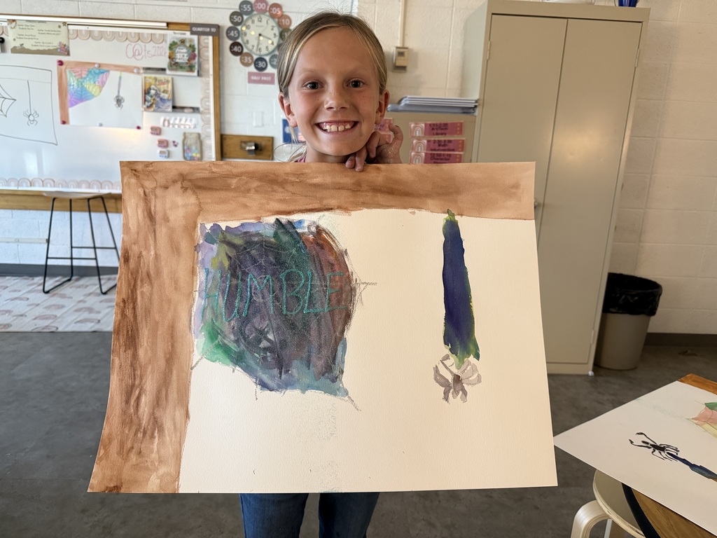 A young student smiling while holding up her colorful watercolor painting of a spiderweb, featuring the word "HUMBLE" written across the center with a spider hanging on the right side.