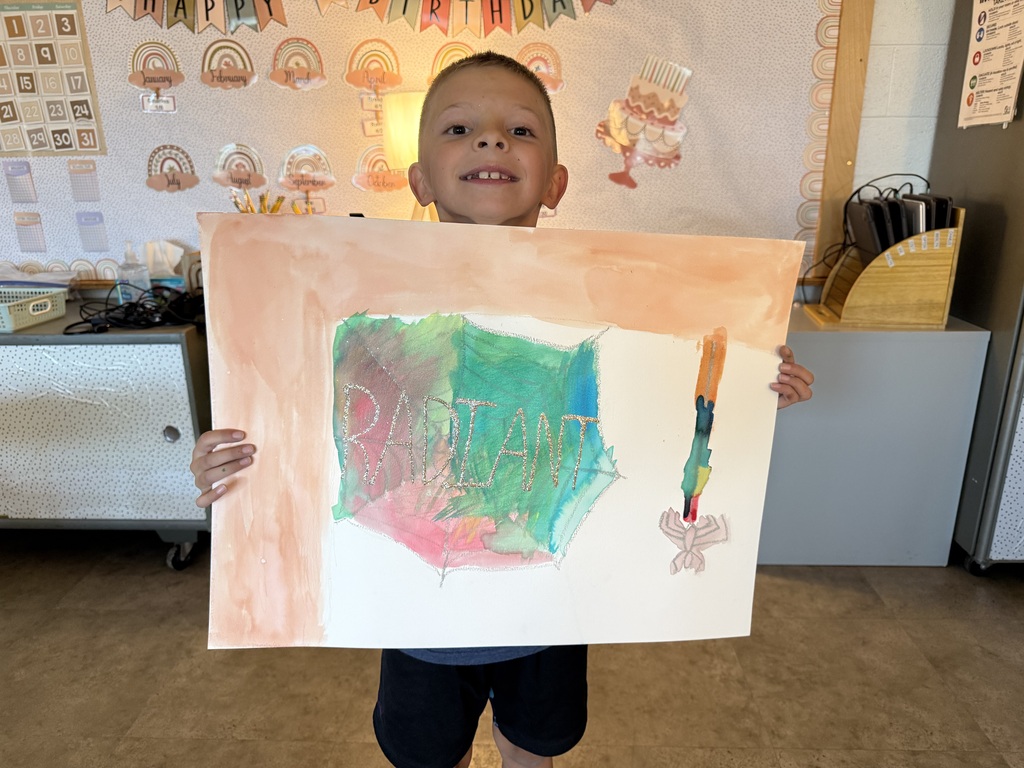 A young boy smiling brightly while holding up his colorful watercolor art project of a spiderweb, featuring the word "RADIANT" written across the center in light-colored lettering with a small spider hanging near the edge.