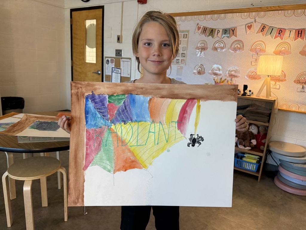 A young student smiling while holding up her colorful watercolor painting of a spiderweb, featuring the word "RADIANT" written in green across the web with a small black spider hanging on the side.