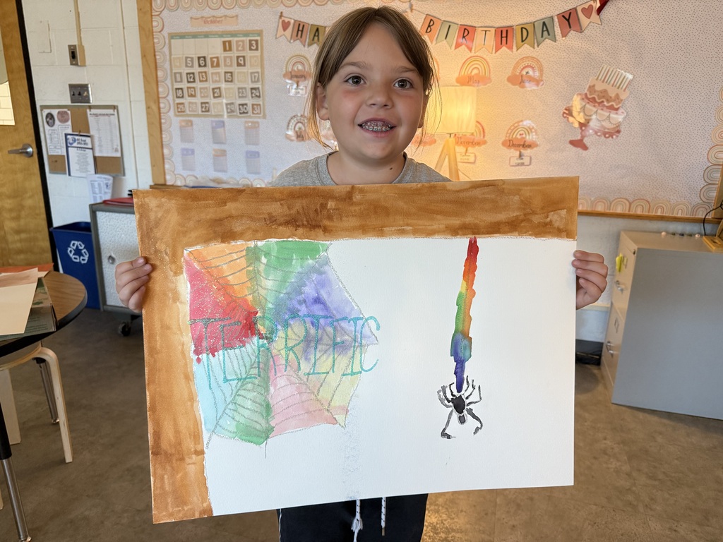 A young student smiling while holding up her colorful watercolor art project of a spiderweb, featuring the word "TERRIFIC" written in blue and green letters with a small spider hanging from a thread on the right.