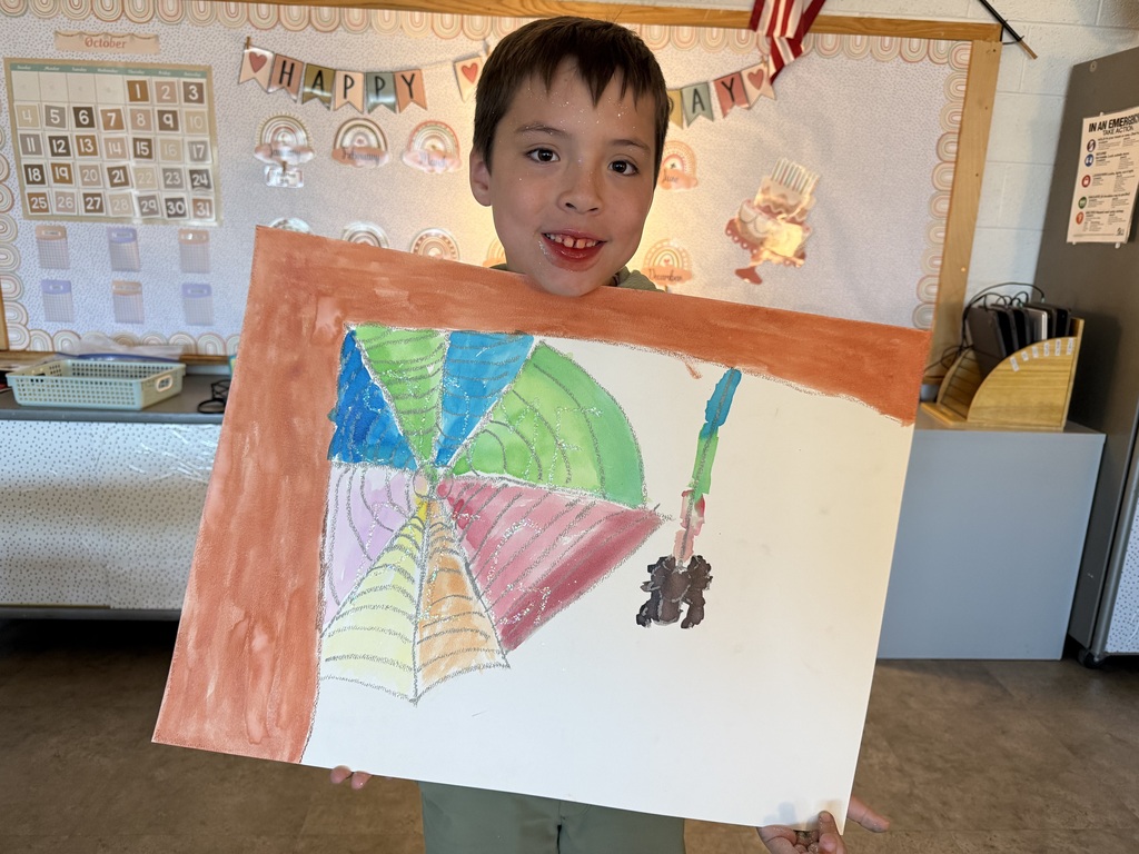 A young boy smiling while holding up his colorful watercolor art project of a spiderweb, featuring a green and orange sectioned web with a small black spider dangling on the right side.