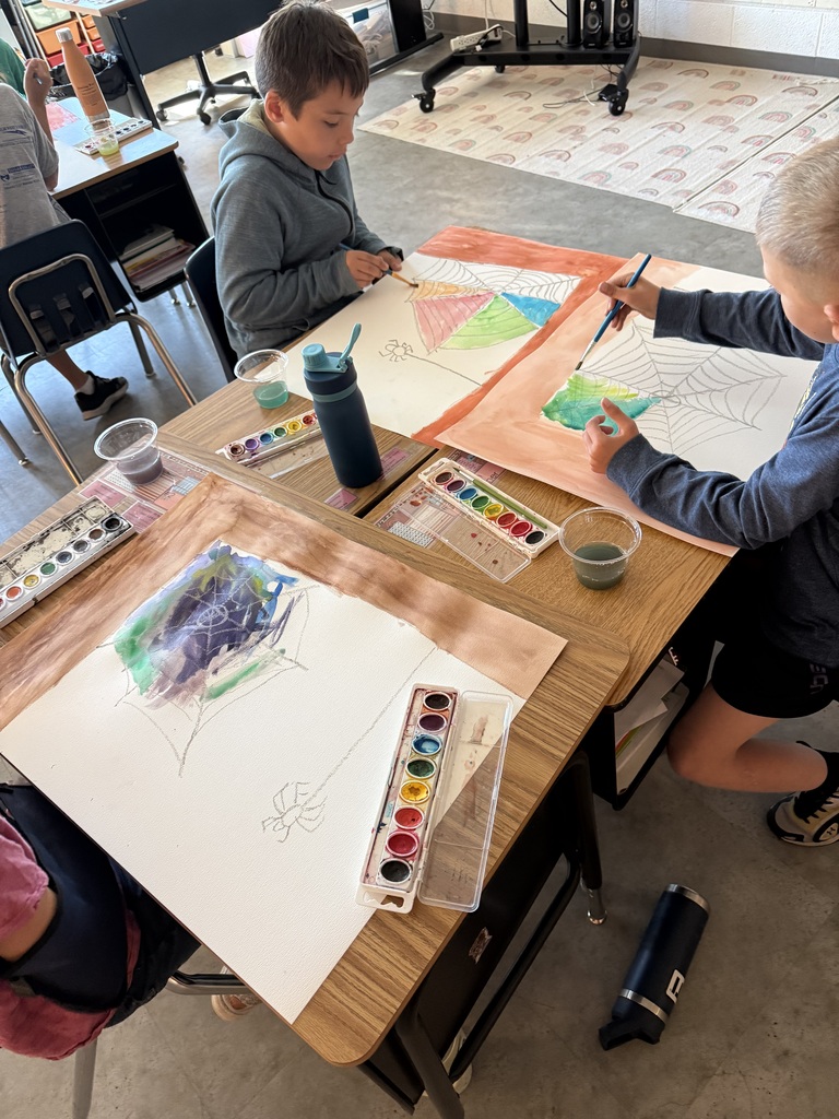 Elementary students sitting at classroom desks working on watercolor spiderweb art projects inspired by Charlotte's Web, using paintbrush trays and water cups.