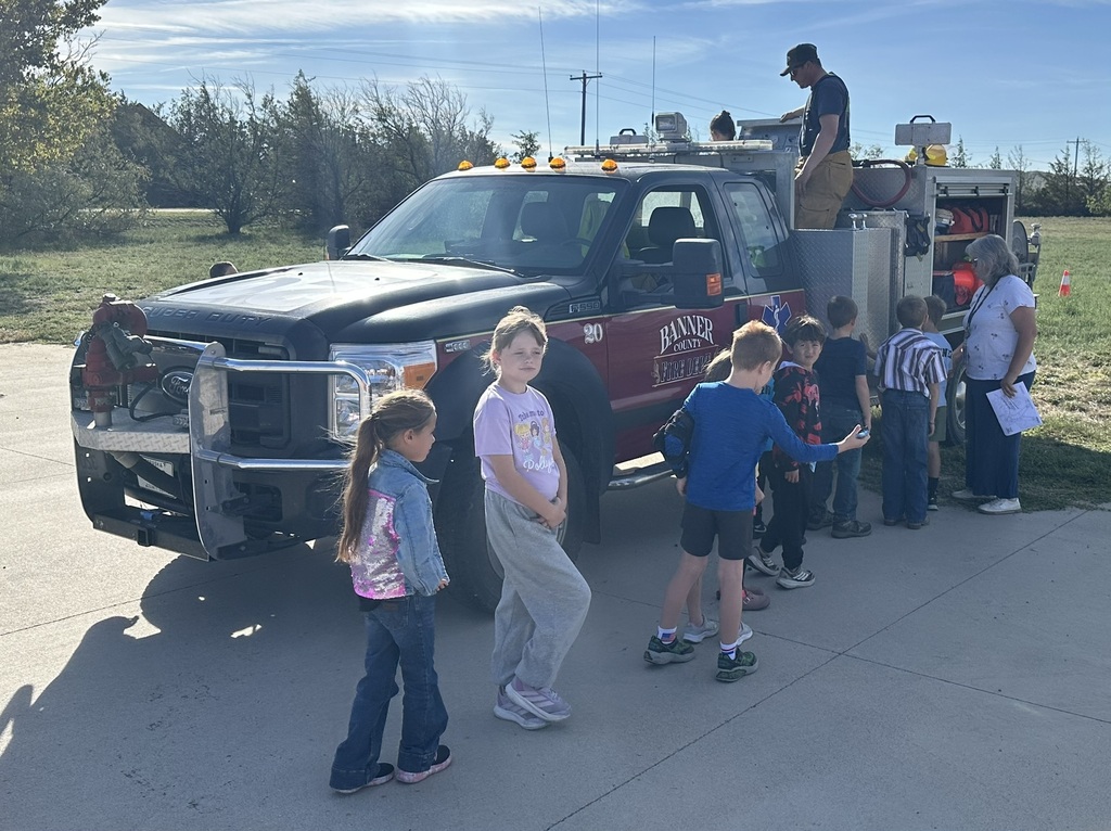 A firefighter in full gear demonstrating equipment to a group of elementary school students standing outside next to a fire truck in front of the school building.
