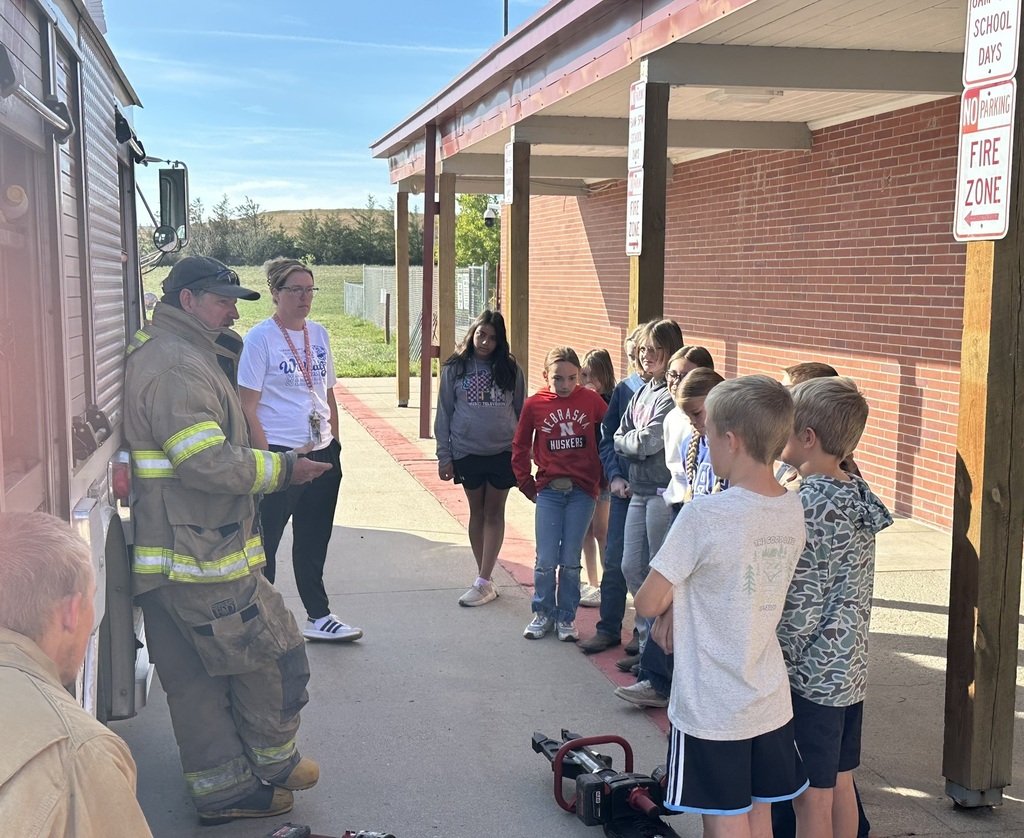 A firefighter in full gear demonstrating equipment to a group of elementary students gathered in a circle outside next to the school building.