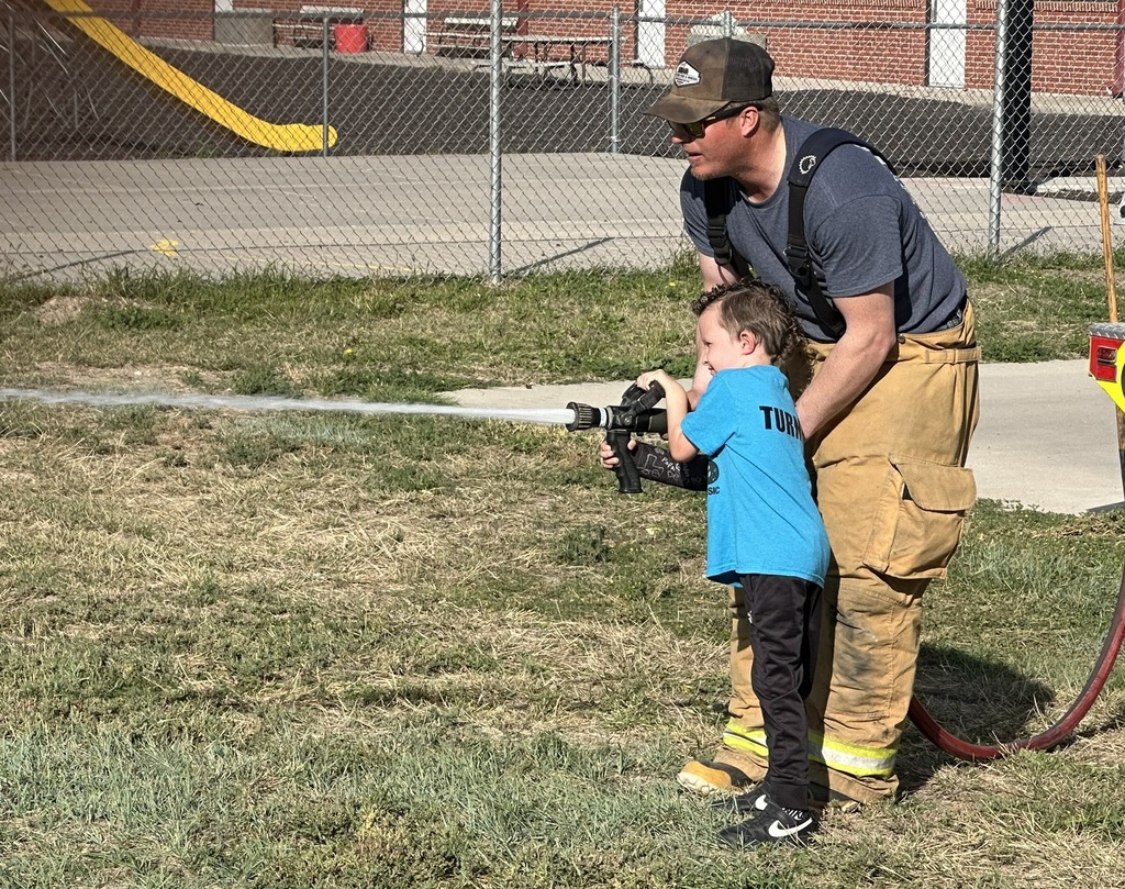A firefighter helping a young boy in a blue shirt hold and spray a fire hose on a grassy area outside the school.