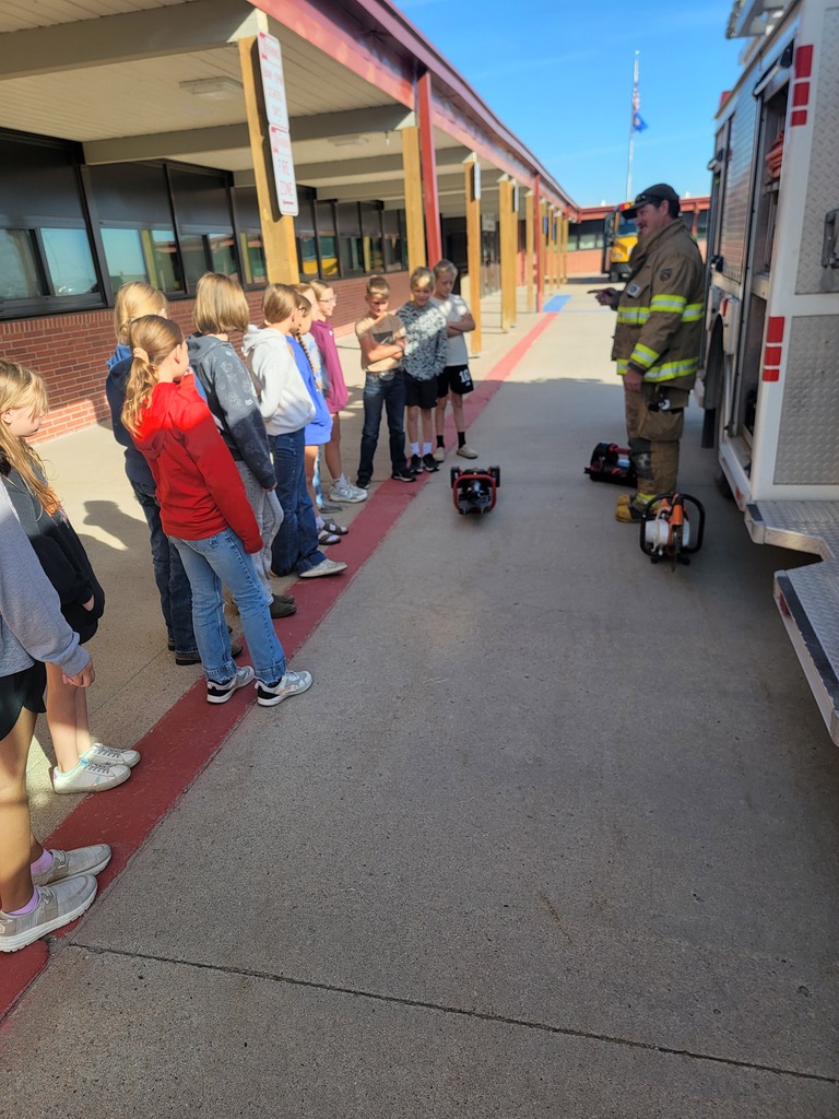 A firefighter in full gear demonstrating equipment to a group of elementary school students standing outside next to a fire truck in front of the school building.