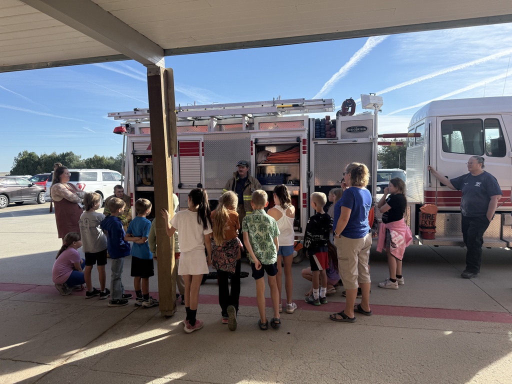 A firefighter in full gear demonstrating equipment to a group of elementary school students standing outside next to a fire truck in front of the school building.