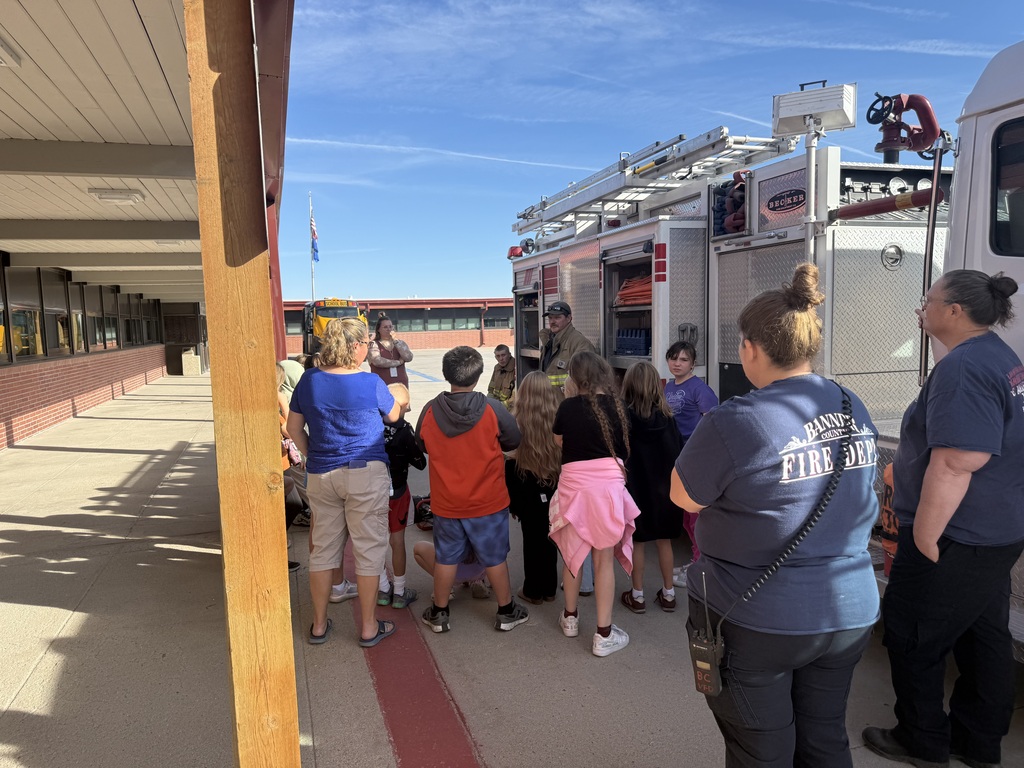 A firefighter in full gear demonstrating equipment to a group of elementary school students standing outside next to a fire truck in front of the school building.