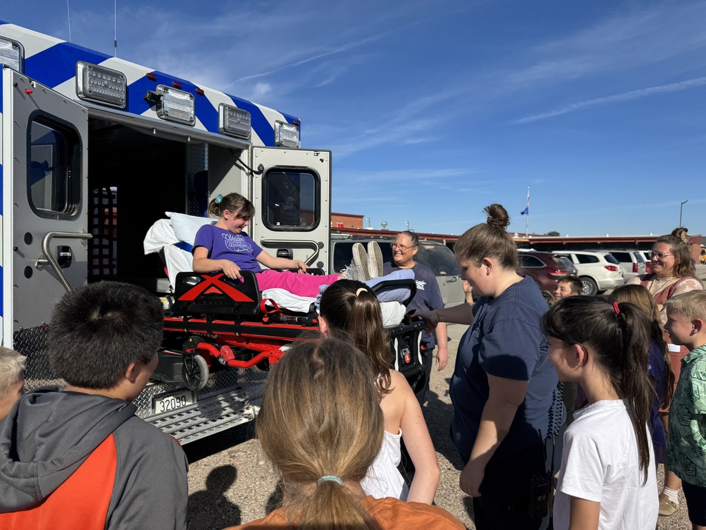 A firefighter in full gear demonstrating equipment to a group of elementary students gathered in a circle outside next to the school building.