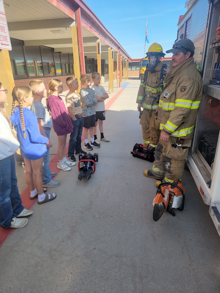 A firefighter in full gear demonstrating equipment to a group of elementary school students standing outside next to a fire truck in front of the school building.