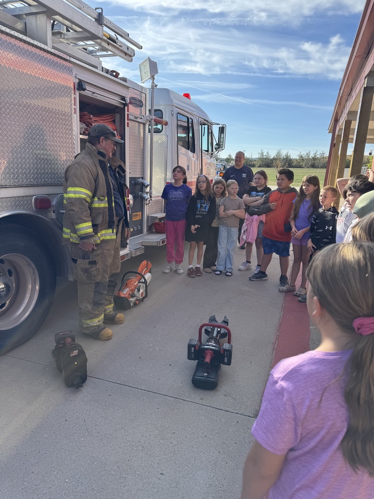 A firefighter in full gear demonstrating equipment to a group of elementary students gathered in a circle outside next to the school building.