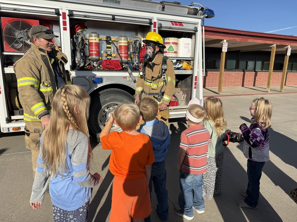 A firefighter in full gear demonstrating equipment to a group of elementary school students standing outside next to a fire truck in front of the school building.