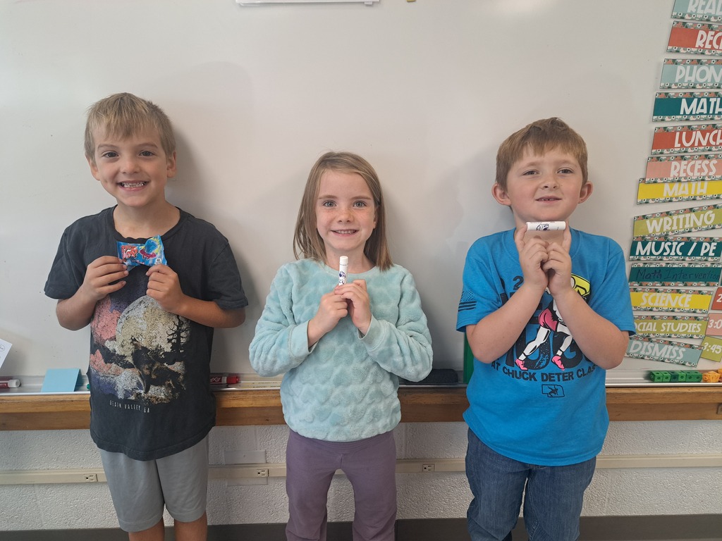 Three elementary students sitting together at a classroom desk, smiling and proudly holding up small items they made or decorated.