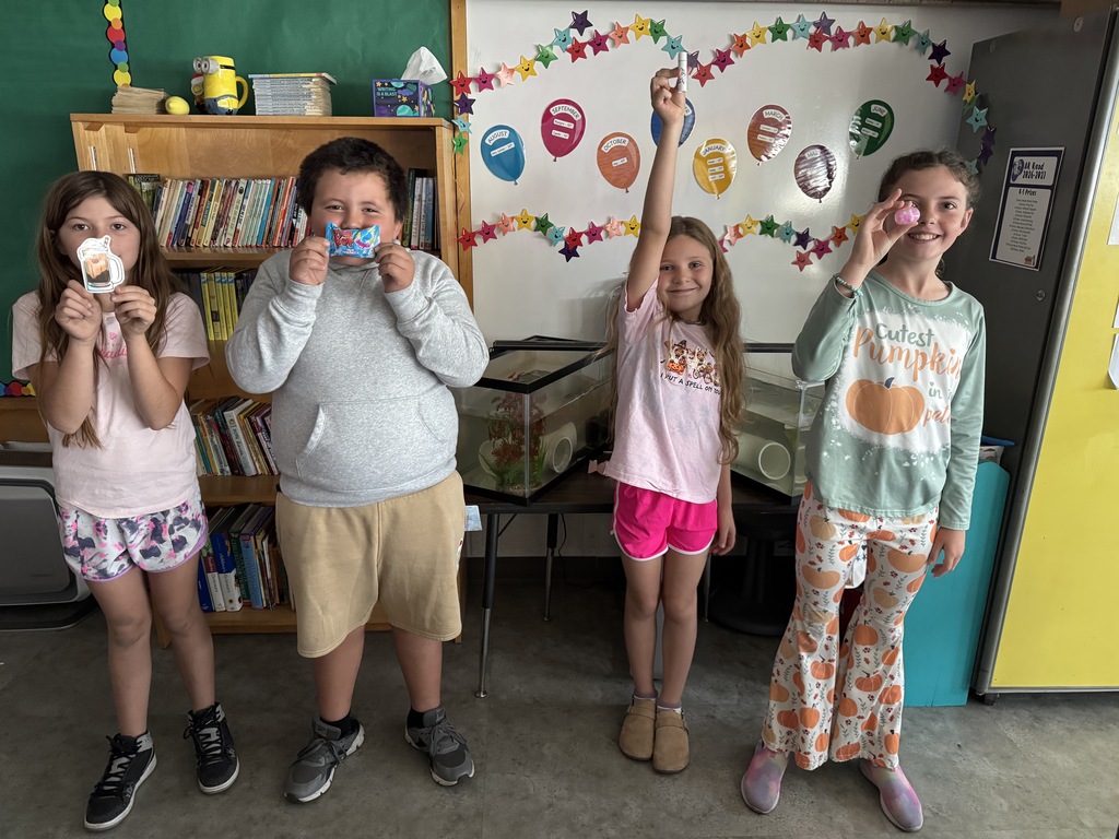 Four elementary students standing in a classroom smiling and holding up small items they made or decorated.
