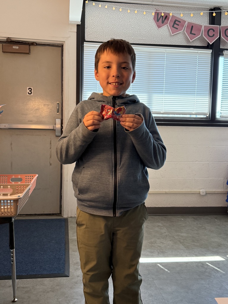 A young boy sitting in a classroom, smiling while holding up a small decorated item or craft in front of a window with a banner behind him.