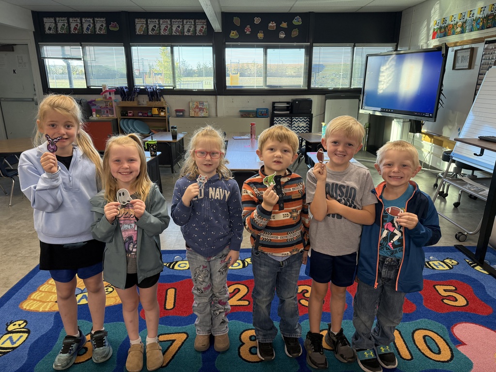 A group of six young elementary students standing together in a classroom, smiling and holding up small handmade crafts.