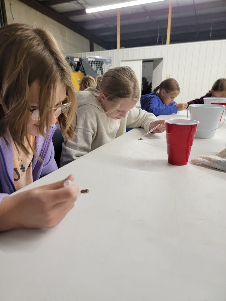 A student in a purple hoodie sitting at a white table, holding a small tool or pipette over a brown spot during a classroom activity.