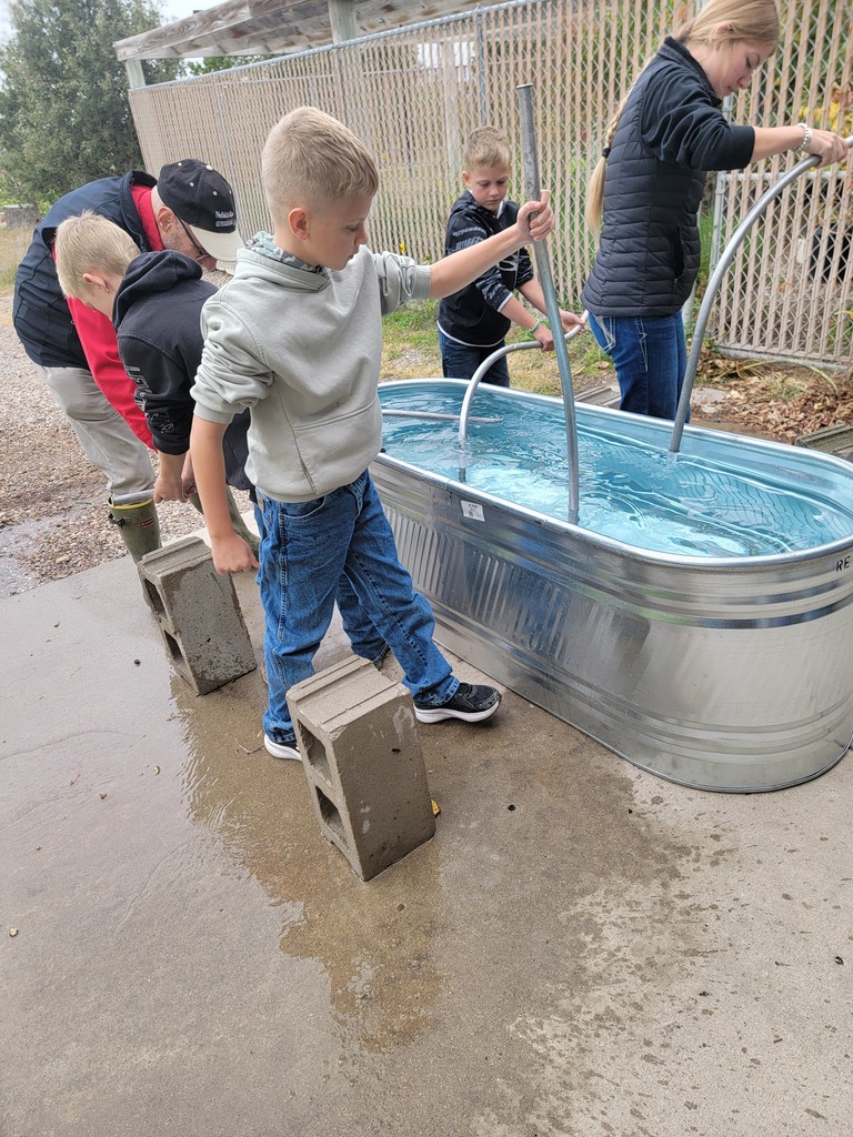 A group of young students outdoors around a large water container, with one student operating a water pump while others lean over to look inside.