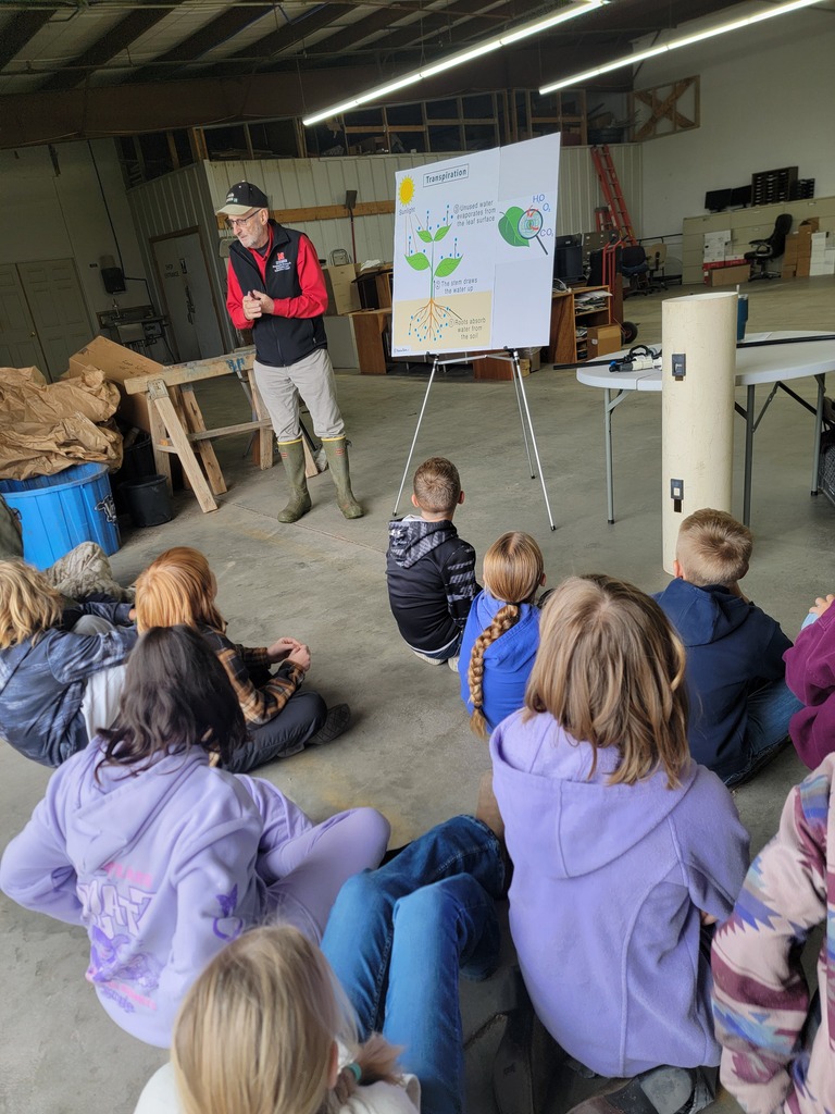 A group of students sitting on the floor in an indoor or sheltered area, listening to a presentation or demonstration.