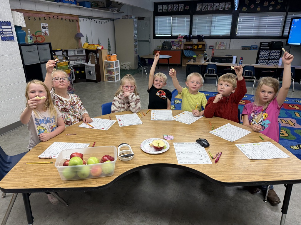 A group of young students sitting around a classroom table during an activity, with several students raising their hands.