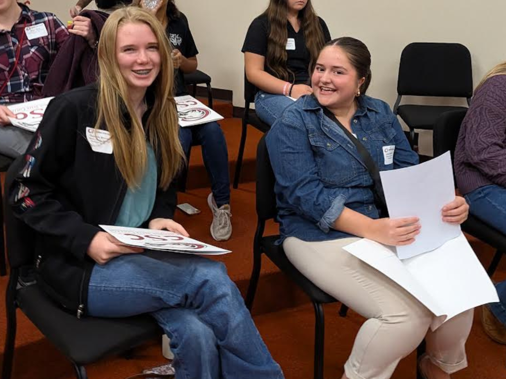 Two female students sitting side-by-side indoors, smiling while holding papers or name tags during an event or presentation.