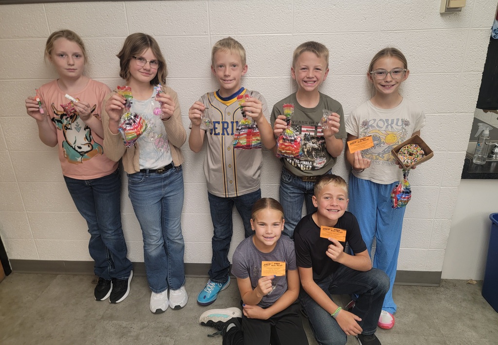 A group of elementary students smiling and posing together indoors while holding small cards or paper badges.
