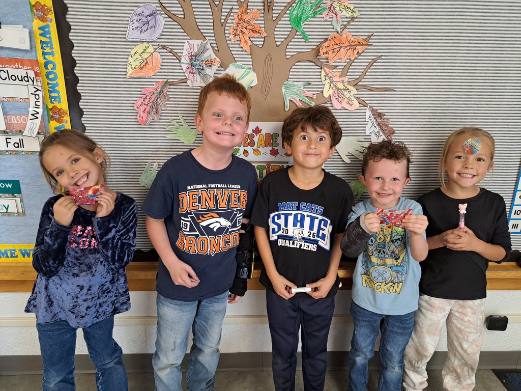 Five young elementary students standing side by side in a classroom, smiling and holding small crafts, in front of a colorful decorated bulletin board.