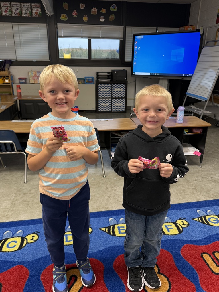 Two young elementary boys standing side-by-side on a colorful alphabet rug in a classroom, smiling while holding small wrapped treats or snacks.