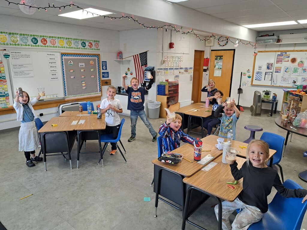 A group of elementary students sitting at their classroom desks, smiling and holding up cups with root beer floats to celebrate.