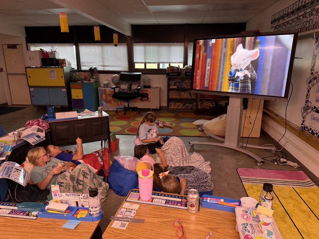 A group of young elementary students relaxing on the floor with blankets and pillows in a classroom, watching a movie or presentation on a large screen display.