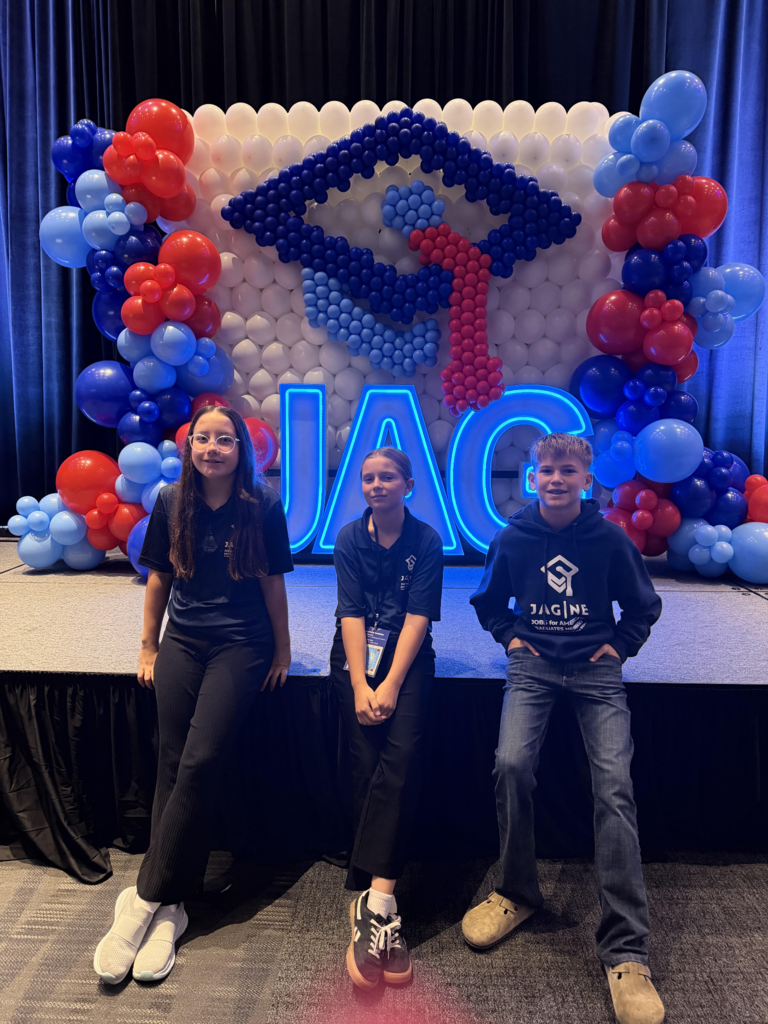 Three JAG students posing on stage in front of a red, white, and blue balloon display surrounding a large JAG logo.