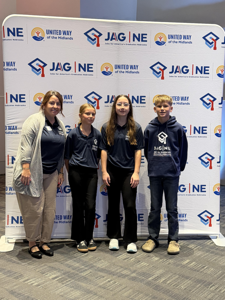 Ms. Villagrana standing with three JAG student officers (Trenton Knaub, Aili Cooper, and Tacey Cross) posing together in front of a JAG Nebraska backdrop.