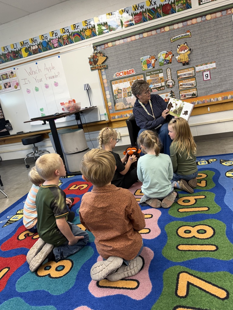 A group of young students sitting on a colorful number rug in a classroom, listening attentively as their teacher reads a book aloud.