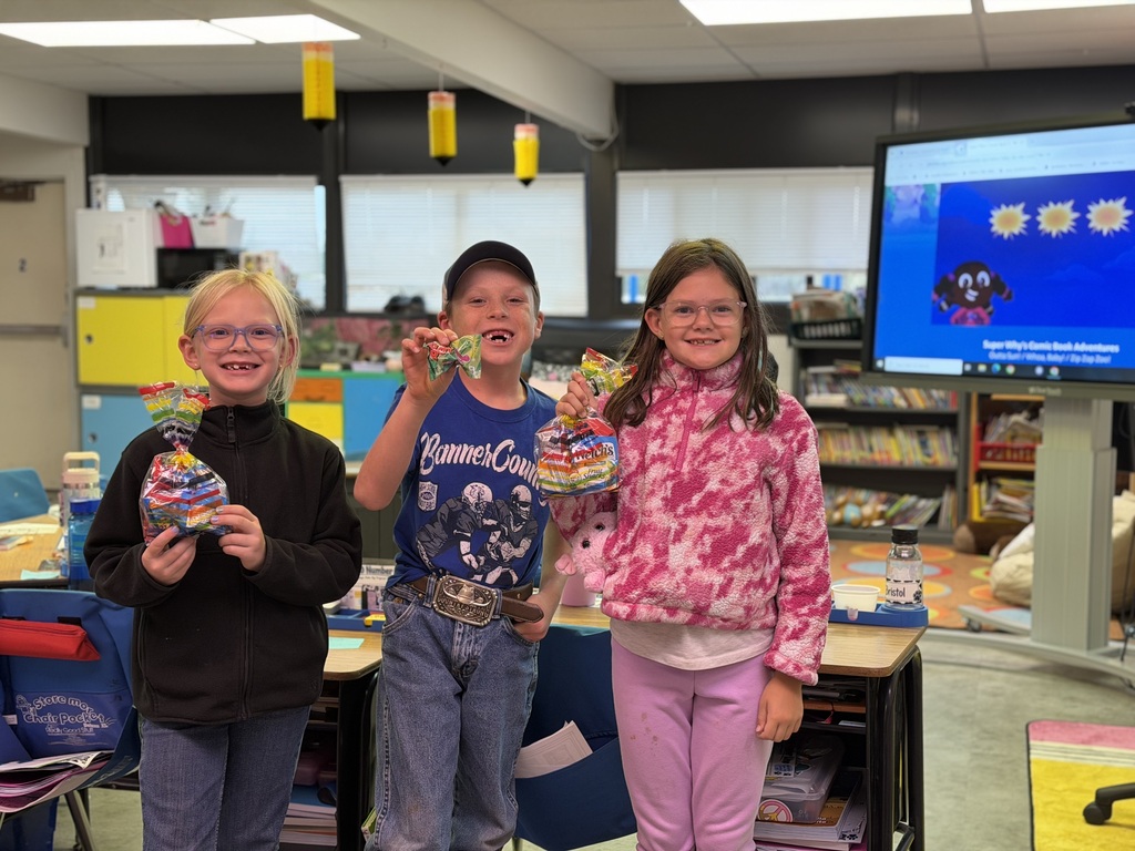 Three young students standing together in a classroom, smiling and holding small craft activities.