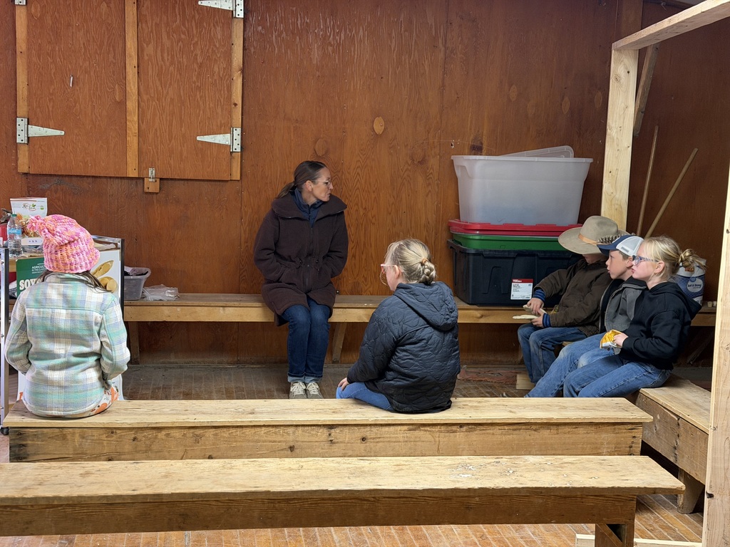 Elementary students sitting on wooden benches inside a picnic shelter during a group presentation.