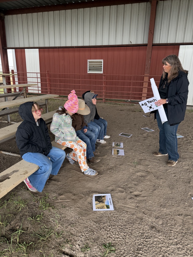 A presenter holding an "Ag Tech" sign while speaking to elementary students sitting on a wooden bench outdoors.