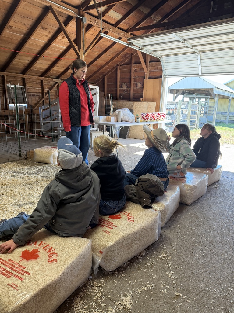 A group of elementary students sitting on hay bales inside a barn, listening to an instructor during a farm field trip.