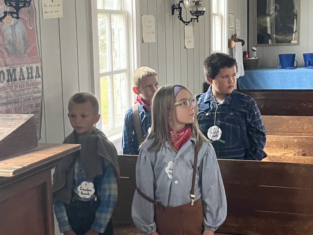 Elementary students in pioneer-style clothing standing near wooden benches inside a historic building.
