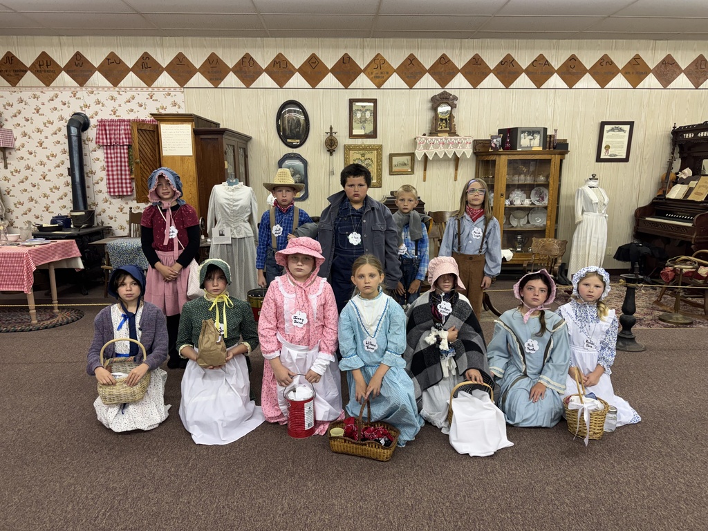 A group of elementary students in pioneer clothing posing together in a museum room with historic artifacts.