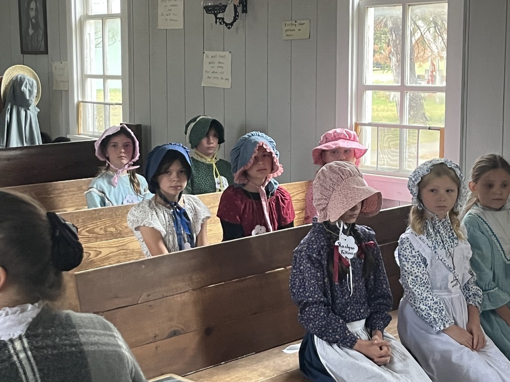 Elementary girls in pioneer dresses and bonnets sitting on wooden pews inside a historic building.