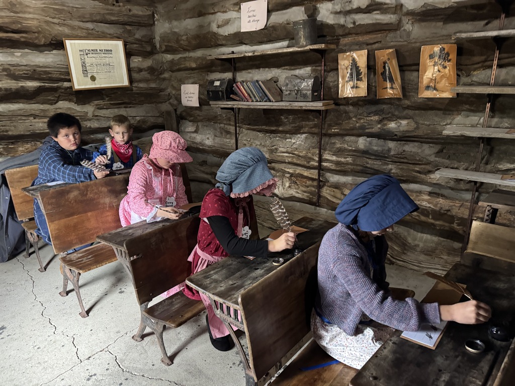 Elementary students in pioneer clothing writing with quill pens at wooden desks inside a historic log cabin classroom.