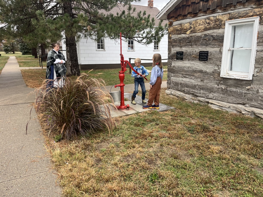 Two elementary students using an antique red hand water pump outside a historic log cabin.