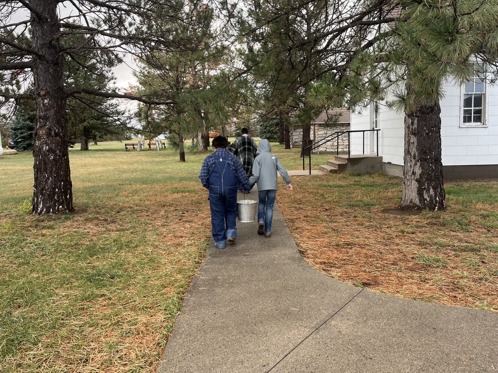 Two elementary students walking down a path while carrying a metal bucket together outside.