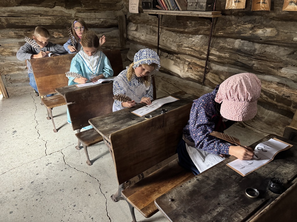 Elementary students in pioneer-style dresses and bonnets writing at wooden desks inside a log cabin.