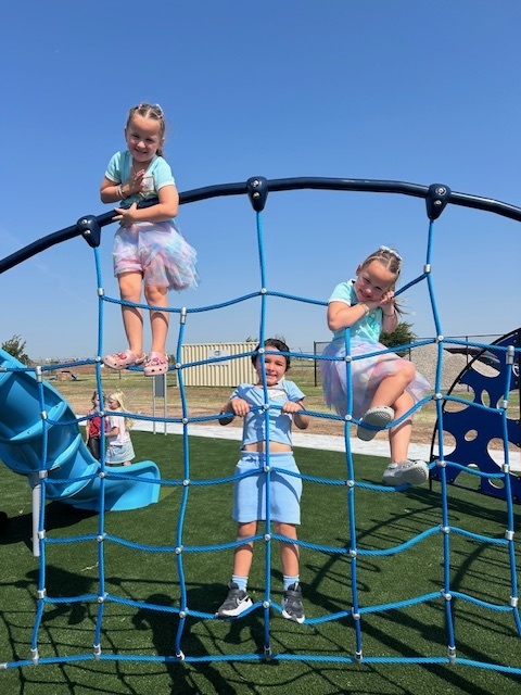 Three children are playing on a blue climbing structure at a playground under a clear blue sky.